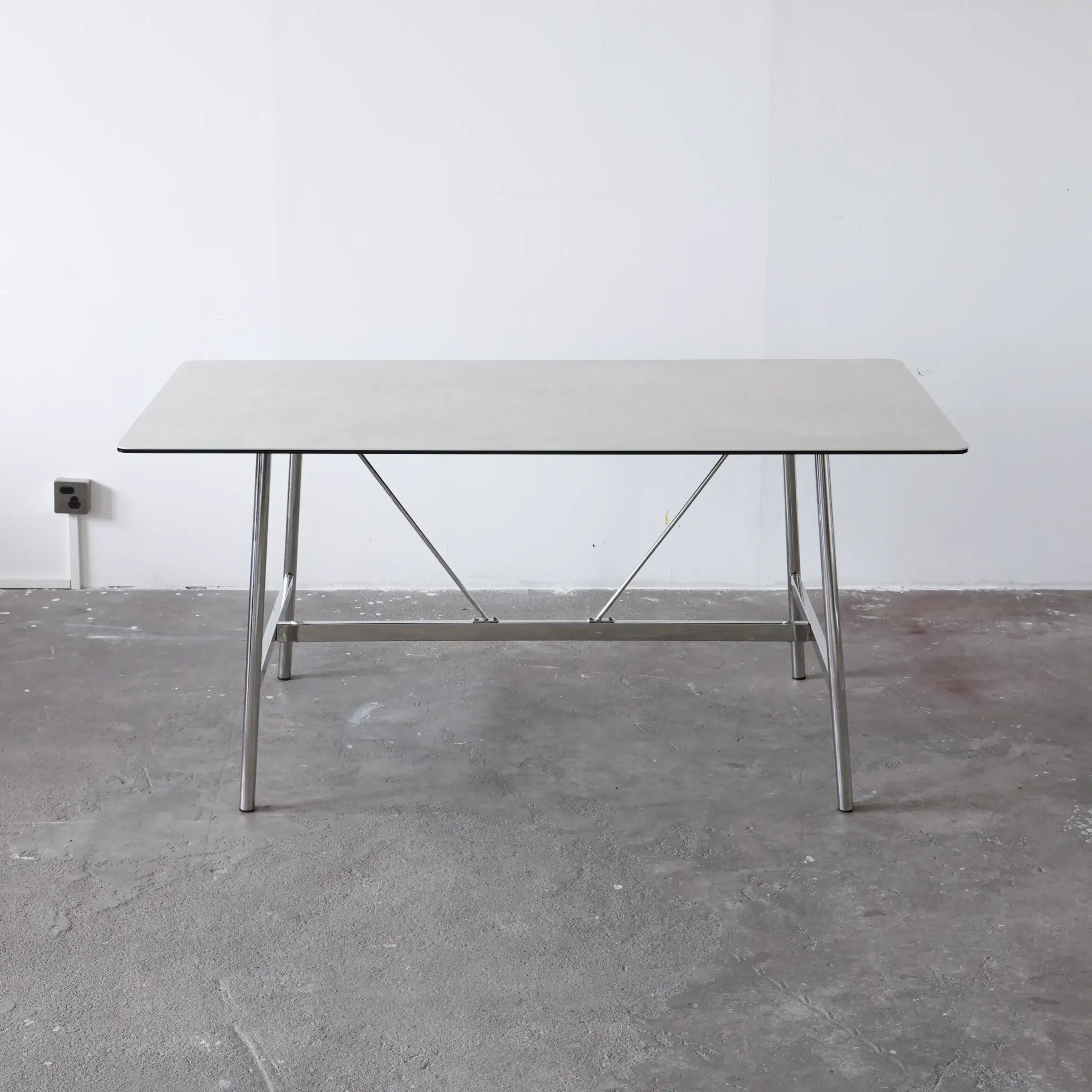A rectangular table with a light-coloured tabletop and metal legs stands on a bare concrete floor against a plain white wall. An electrical socket is visible on the left side of the wall.