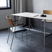 A minimalist office setup with a table, two chairs, a book, and black headphones on a tiled floor near a window with light grey curtains.
