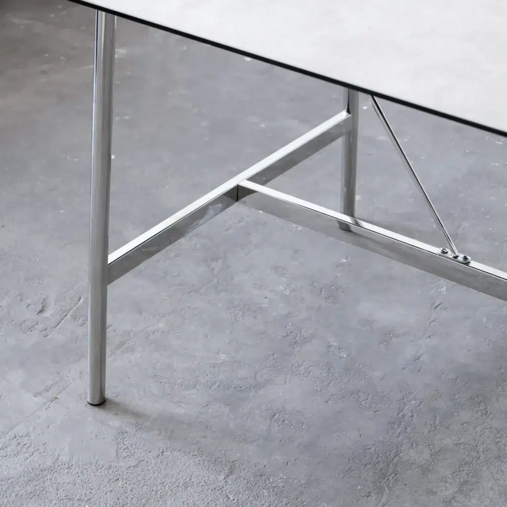 Close-up of a modern table with a thin, light-coloured top and shiny metal legs on a grey, textured floor. The table has a minimalist design with visible support bars under the surface.