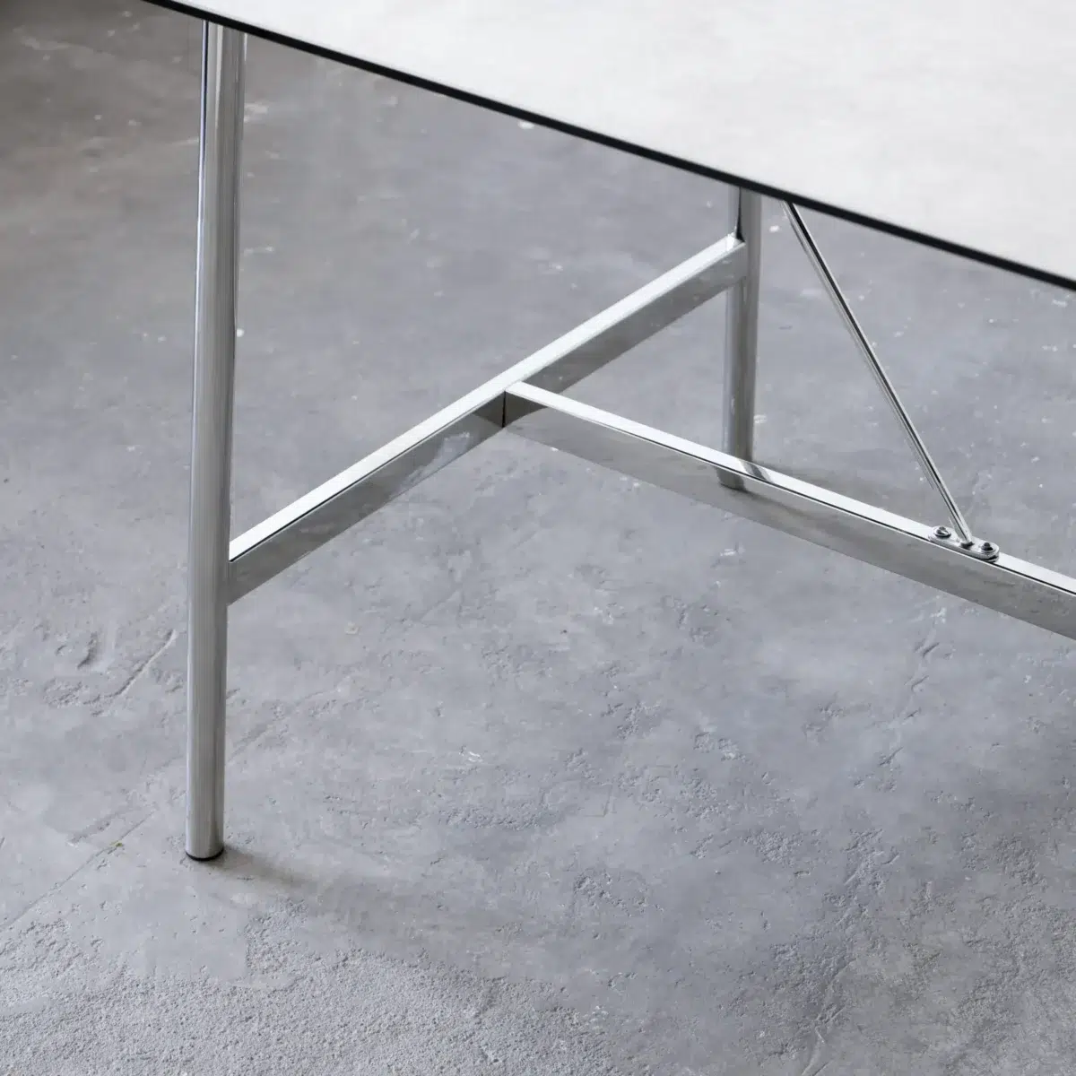 Shuge Dining Table_White_Front View_Detail(2) Close-up of a modern table with a thin, light-coloured top and shiny metal legs on a grey, textured floor. The table has a minimalist design with visible support bars under the surface.