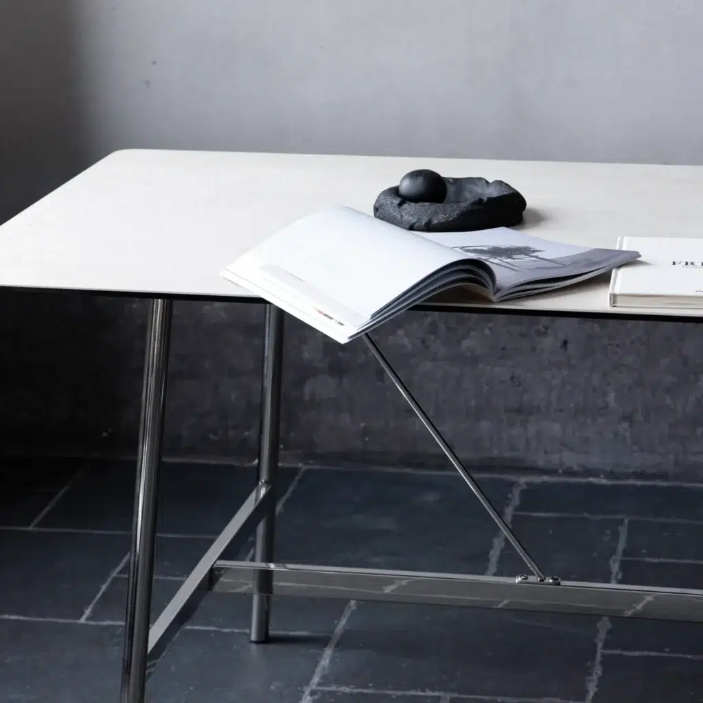 A modern table with metal legs holds an open book, a closed book, and a decorative black stone sculpture on a rectangular ceramic tray, set against a grey wall and dark tiled floor.