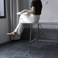 A person in a black shirt and white trousers sits on the edge of a table, holding a book with legs crossed. The room has grey walls, tiled floor, and a window with light curtains.