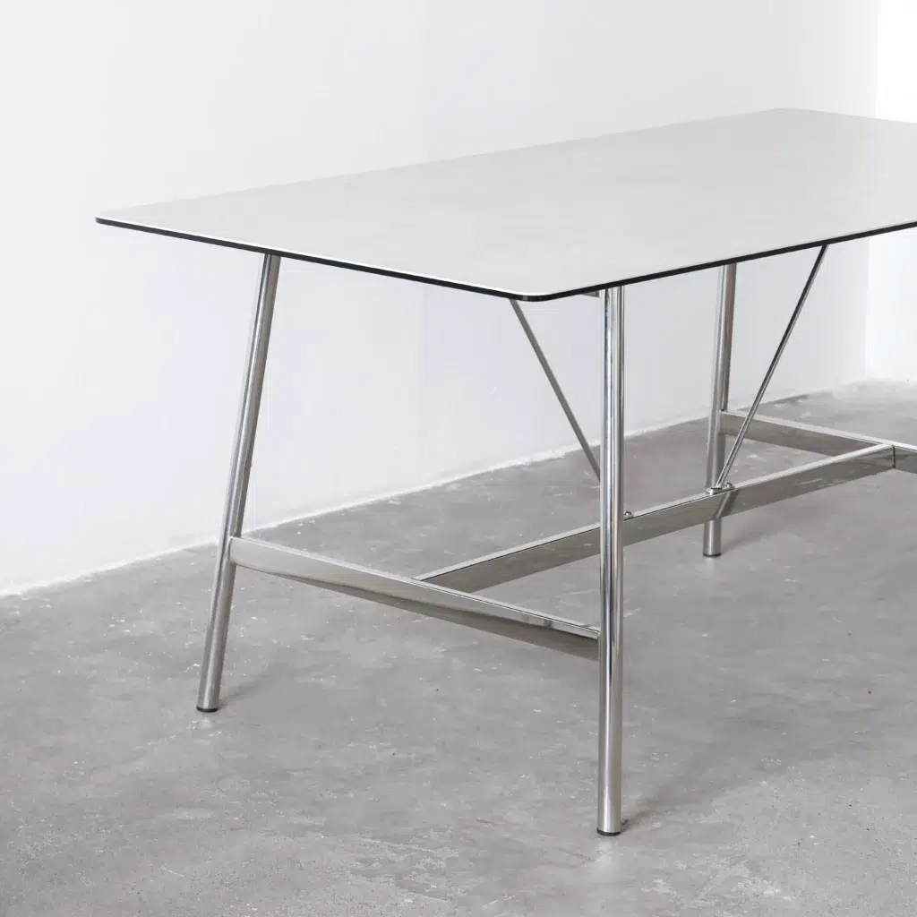 A minimalist rectangular table with a thin, light-coloured tabletop and metal legs, set on a plain grey floor against a white wall.