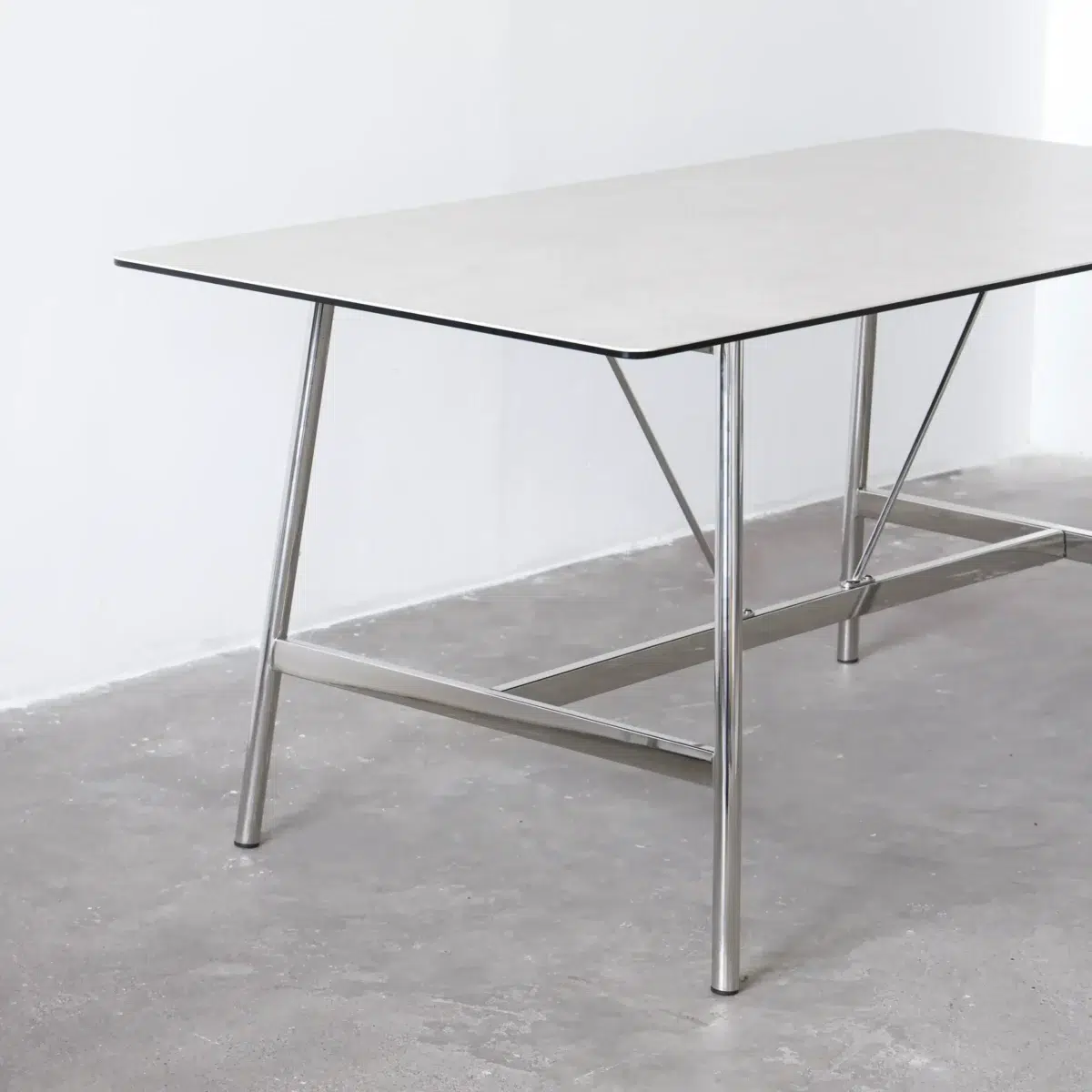Shuge Dining Table_White_Side View A minimalist rectangular table with a thin, light-coloured tabletop and metal legs, set on a plain grey floor against a white wall.