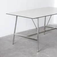 A minimalist rectangular table with a thin, light-coloured tabletop and metal legs, set on a plain grey floor against a white wall.