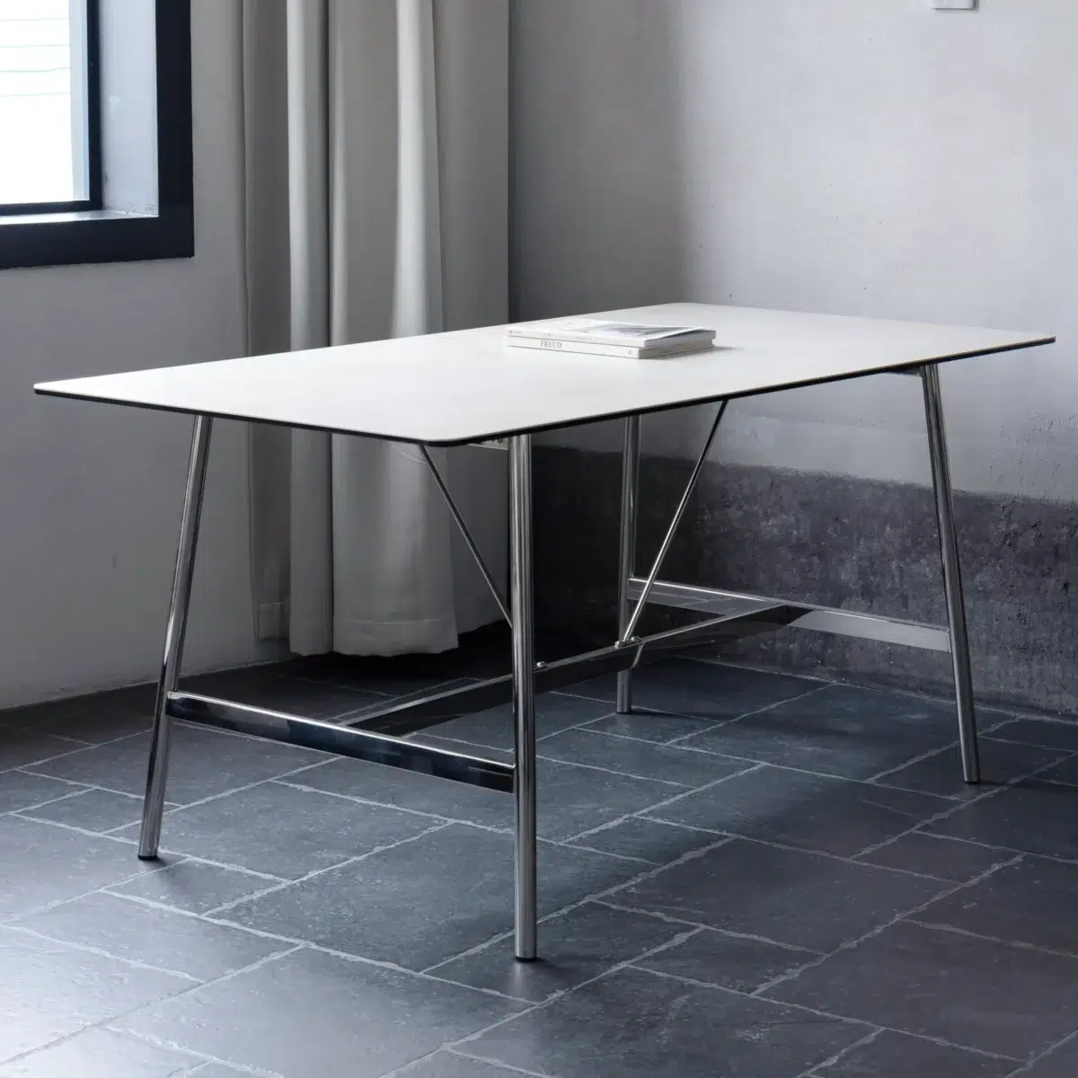 Shuge Dining Table_White_Side View(2) A modern, minimalist table with a thin white tabletop and metal legs stands on a dark tiled floor near a window with white curtains. A closed book is placed at the centre of the table.