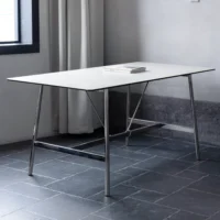 A modern, minimalist table with a thin white tabletop and metal legs stands on a dark tiled floor near a window with white curtains. A closed book is placed at the centre of the table.