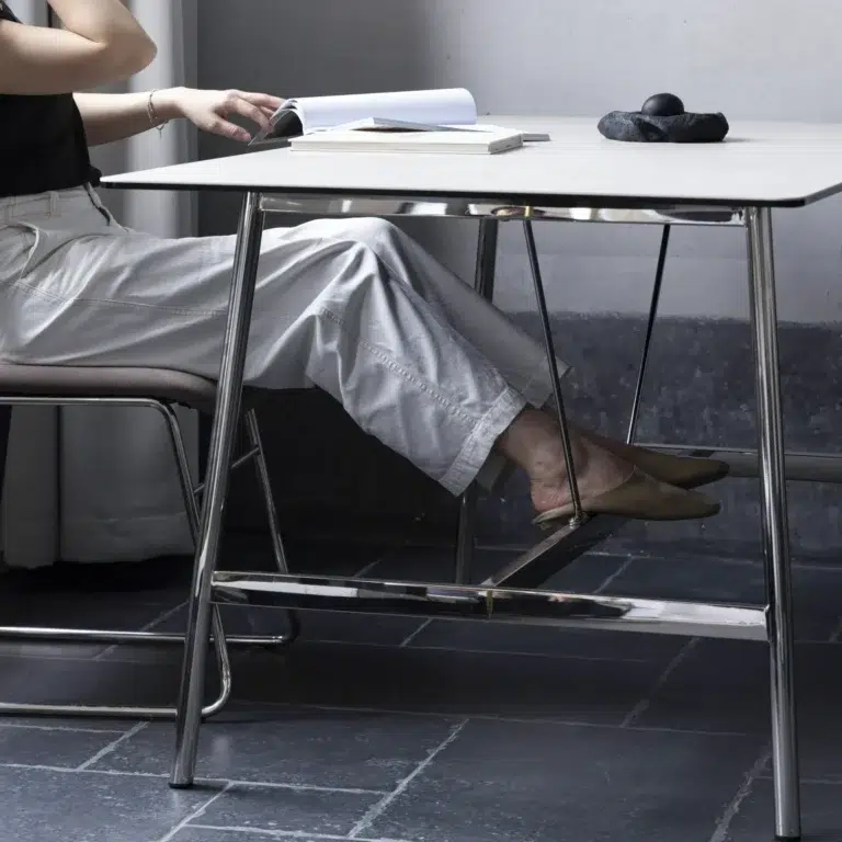 A person in white trousers and tan shoes sits at a modern desk with metal legs, writing in a notebook. Only their lower body and hand are visible. The floor is tiled, and there are small objects on the table.