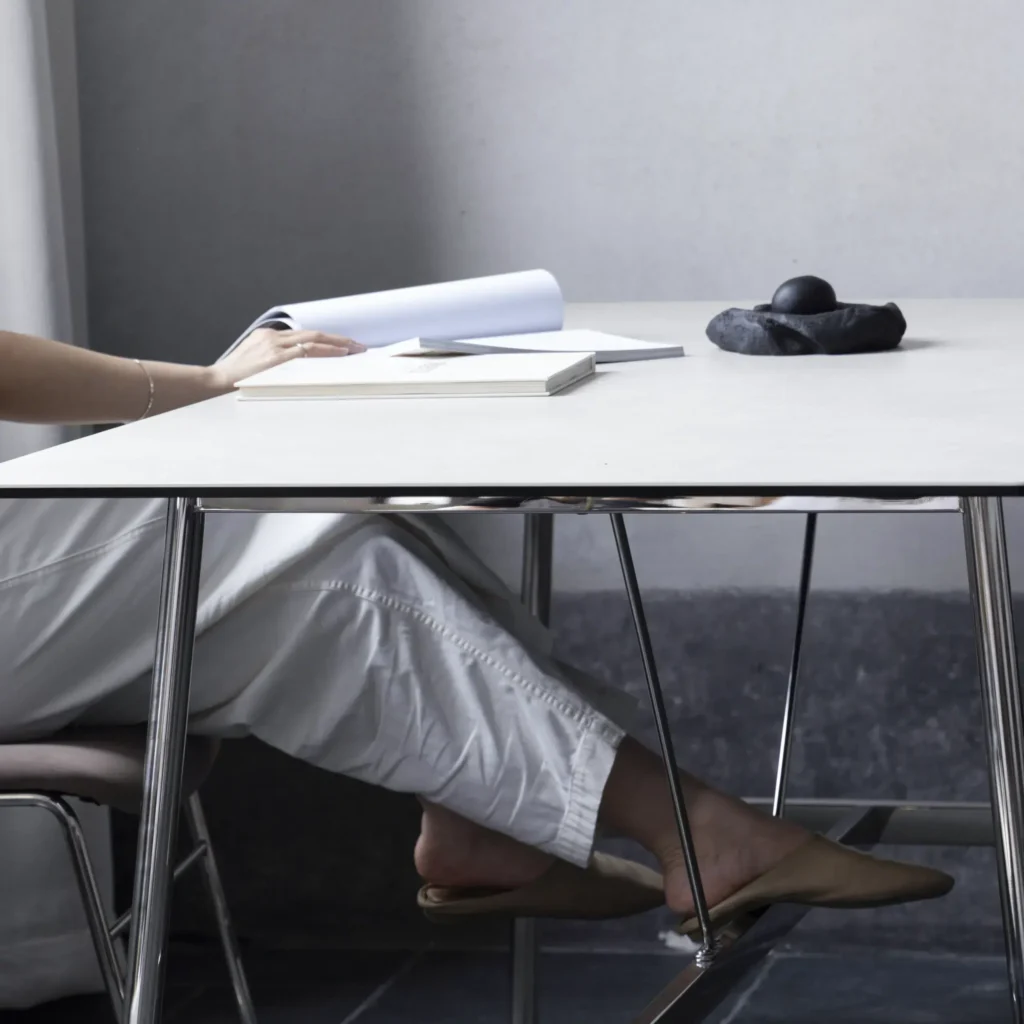 A person dressed in white clothing sits at a minimalist desk with chrome legs, reading a book. Other books and an artistic black stone sculpture are on the table. Only the person’s arm and legs are visible.