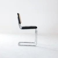 A modern cantilever chair with a chrome metal frame, black seat, and woven cane backrest, displayed in profile against a plain white background.