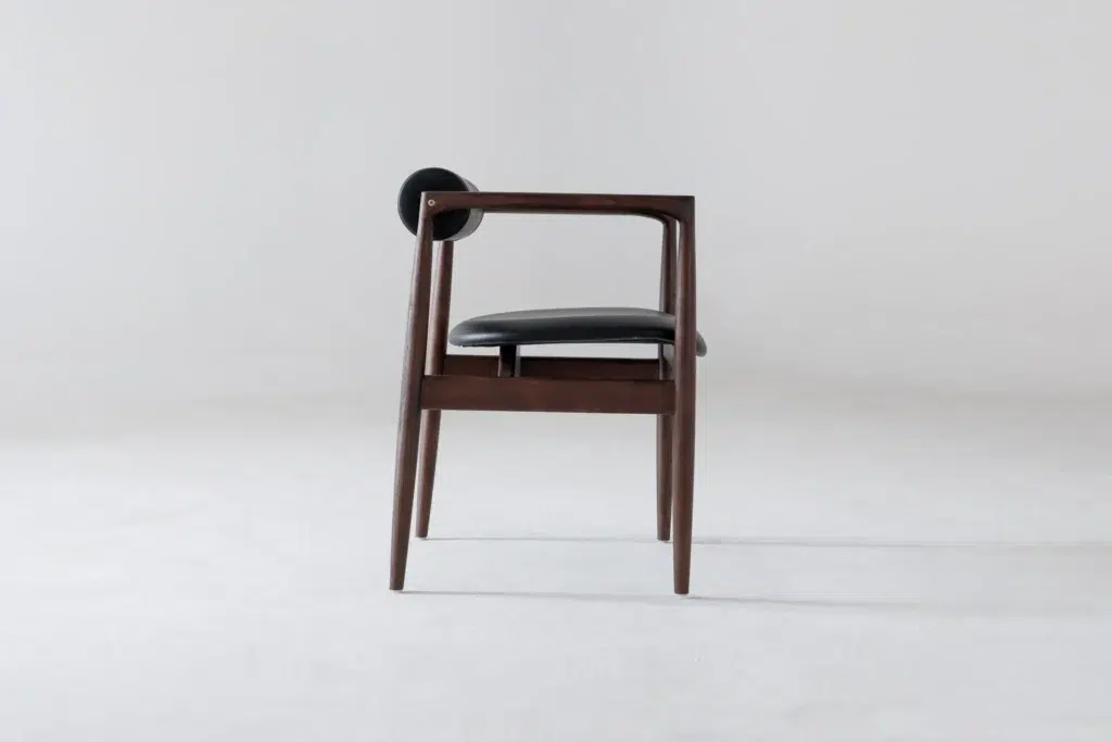 A modern wooden chair with a dark finish, black cushioned seat, and a round black backrest, positioned sideways against a plain light grey background.