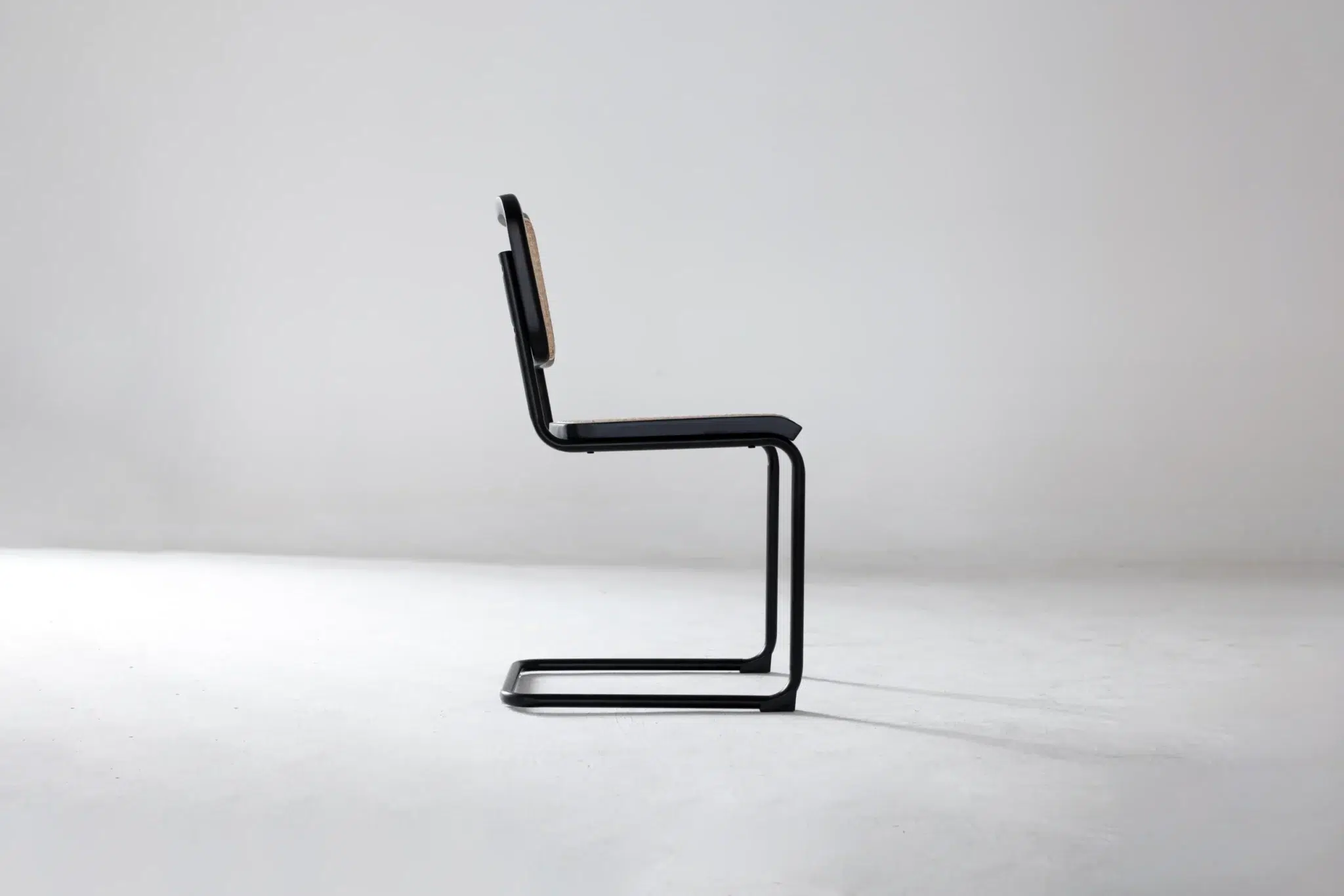 A modern, minimalist chair with a black metal cantilever frame and thin seat and backrest, photographed in profile against a plain, light grey background.