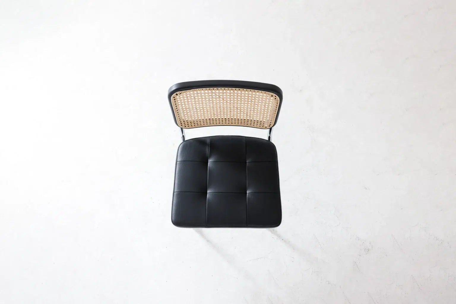 Top view A top-down view of a chair with a woven cane backrest and a black, tufted leather seat, placed on a light, textured surface.