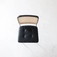 A top-down view of a chair with a woven cane backrest and a black, tufted leather seat, placed on a light, textured surface.