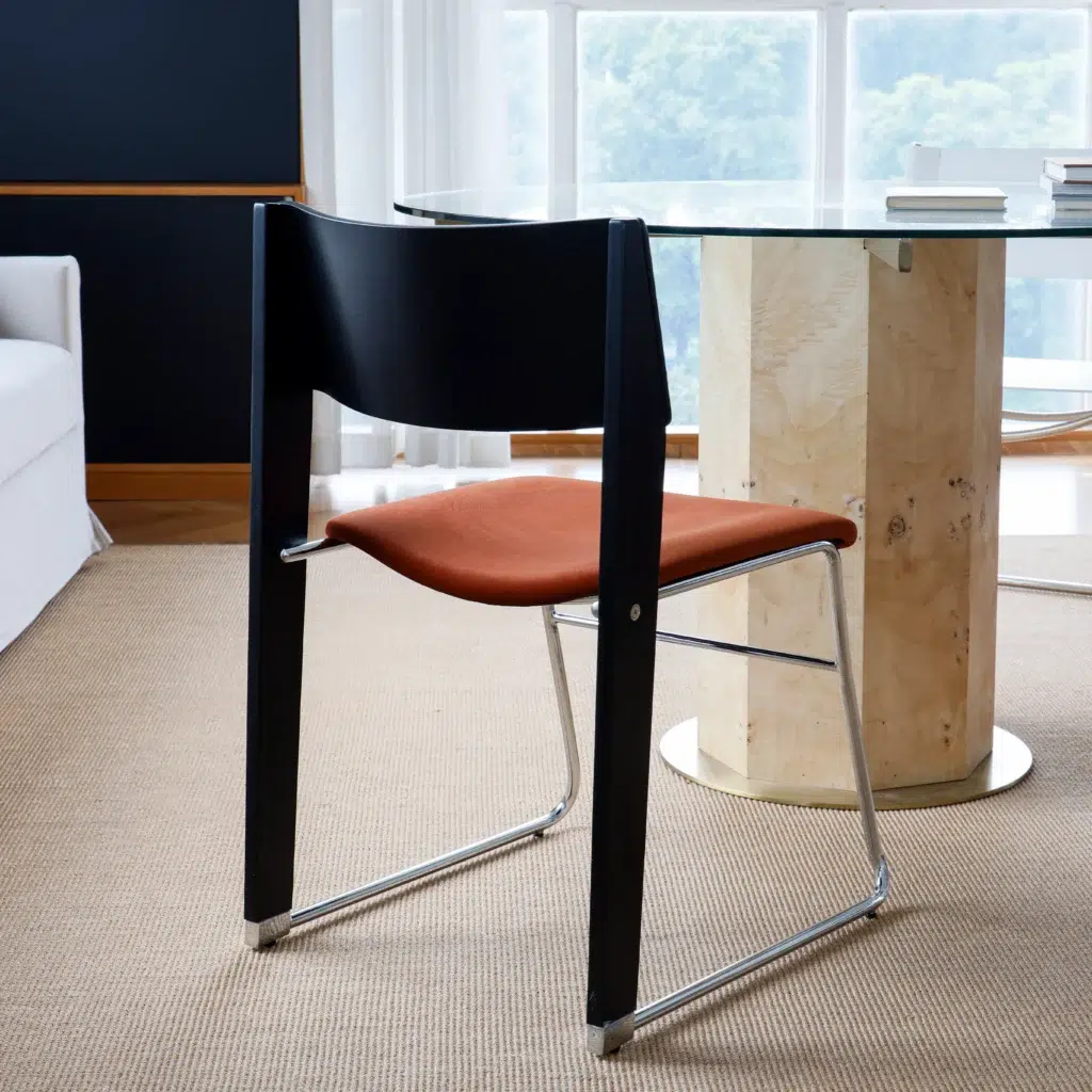 A modern chair with a black backrest, rust-coloured seat, and metal legs is placed next to a round glass table with a light wooden base in a bright room with large windows and beige carpet.