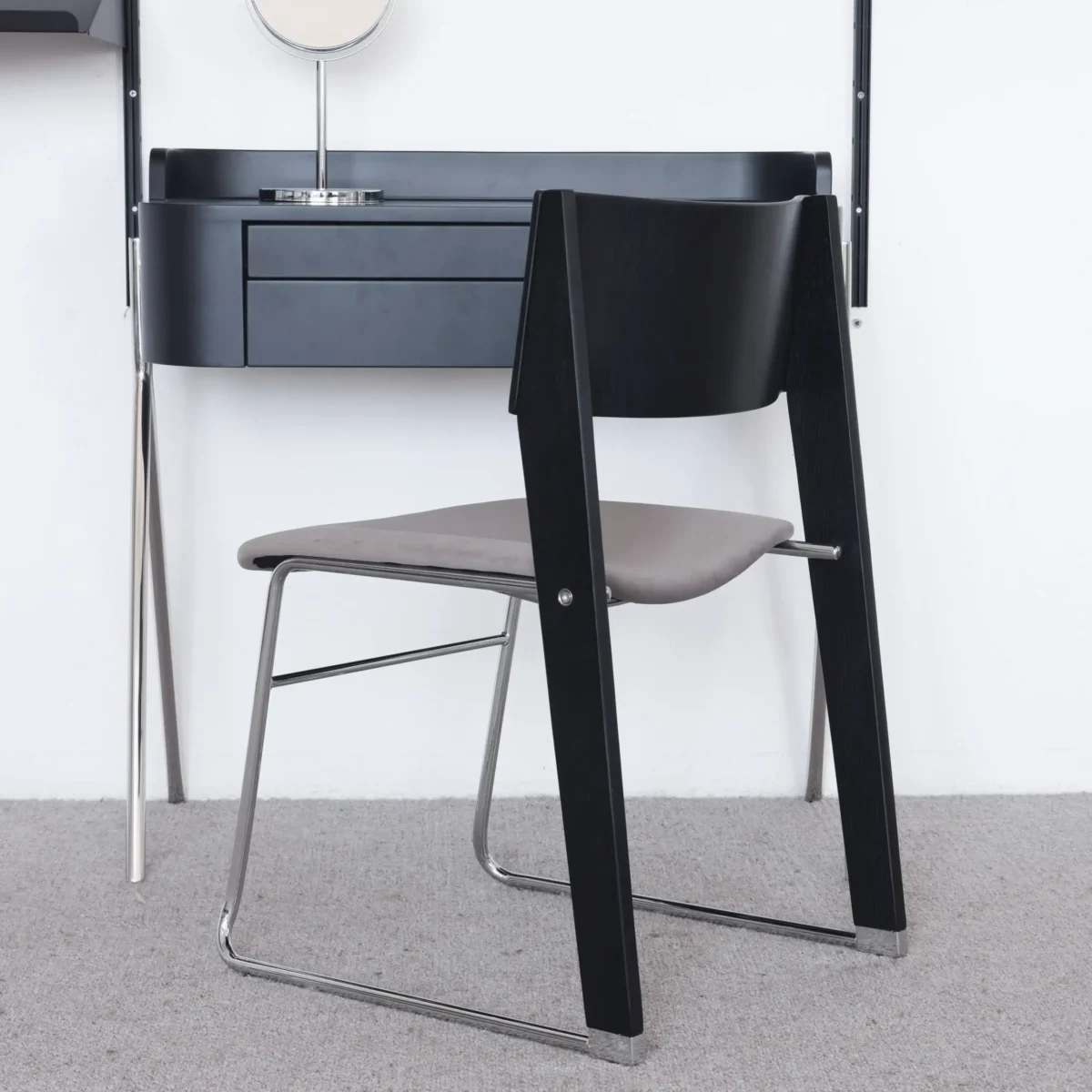 Walk Slowly Chair_Black_Back View(6) A modern chair with a black wooden backrest, grey seat, and chrome legs sits in front of a minimalist black desk with drawers, on a light grey carpeted floor.