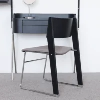 A modern chair with a black wooden backrest, grey seat, and chrome legs sits in front of a minimalist black desk with drawers, on a light grey carpeted floor.