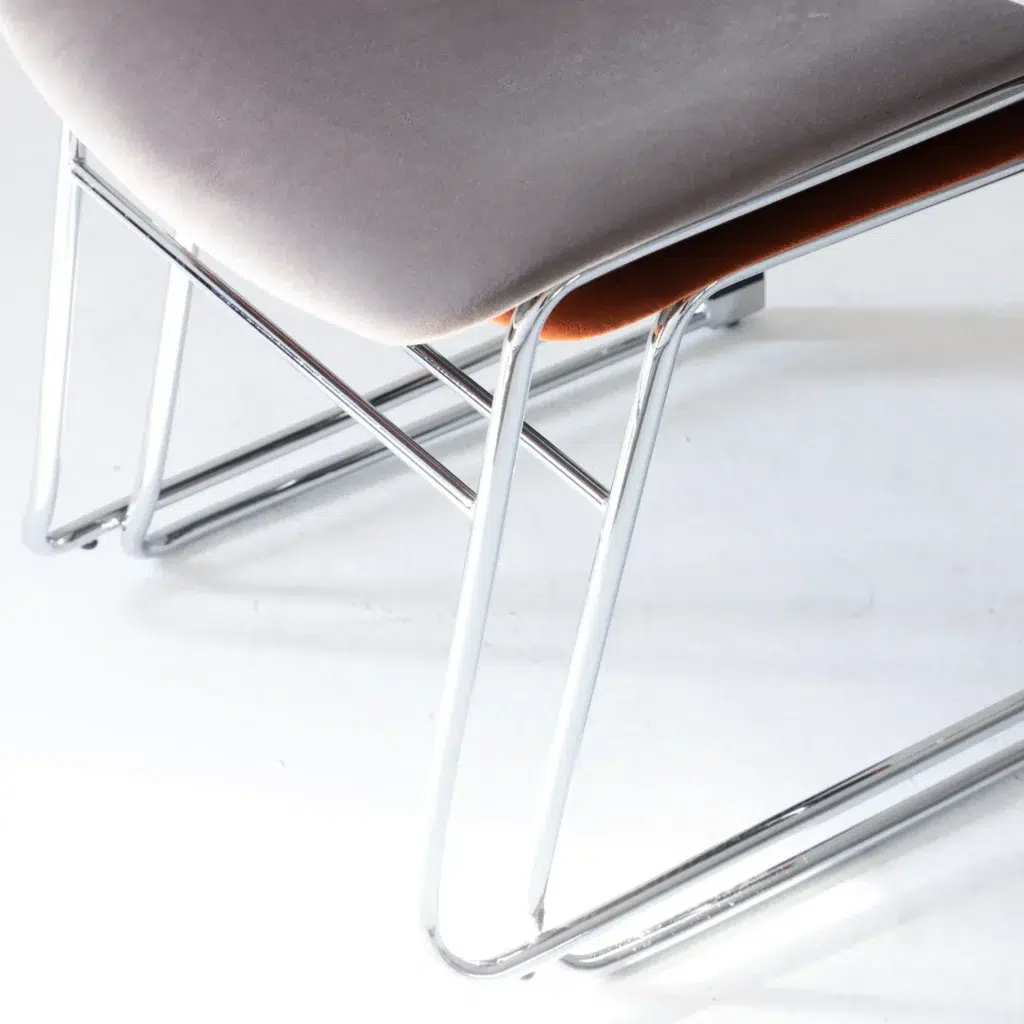 A close-up of a modern chair with a light grey cushioned seat and shiny chrome metal legs, positioned on a white floor. The image focuses on the lower part of the chair and its sleek design.