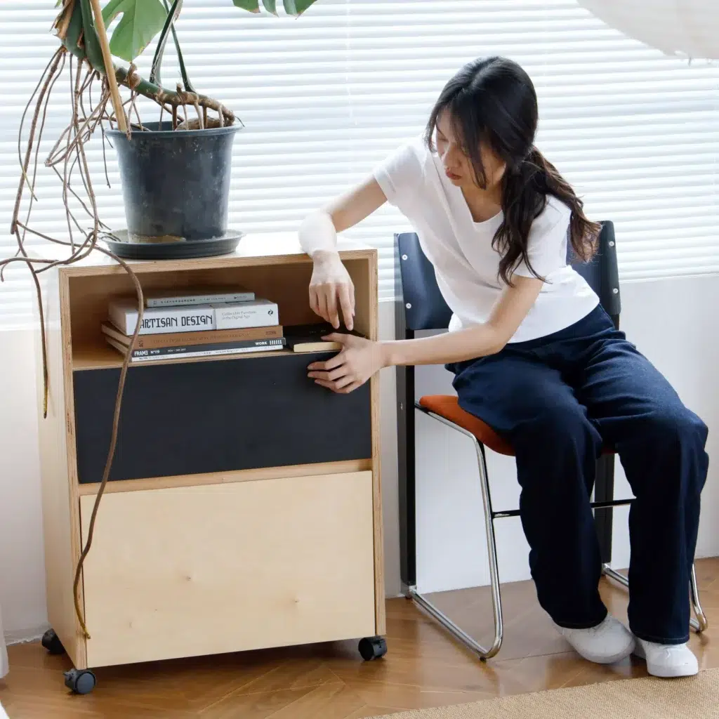A woman in a white T-shirt and dark trousers sits on a chair next to a small rolling wooden cabinet, placing a book on a shelf beneath a potted plant in a bright room with blinds.