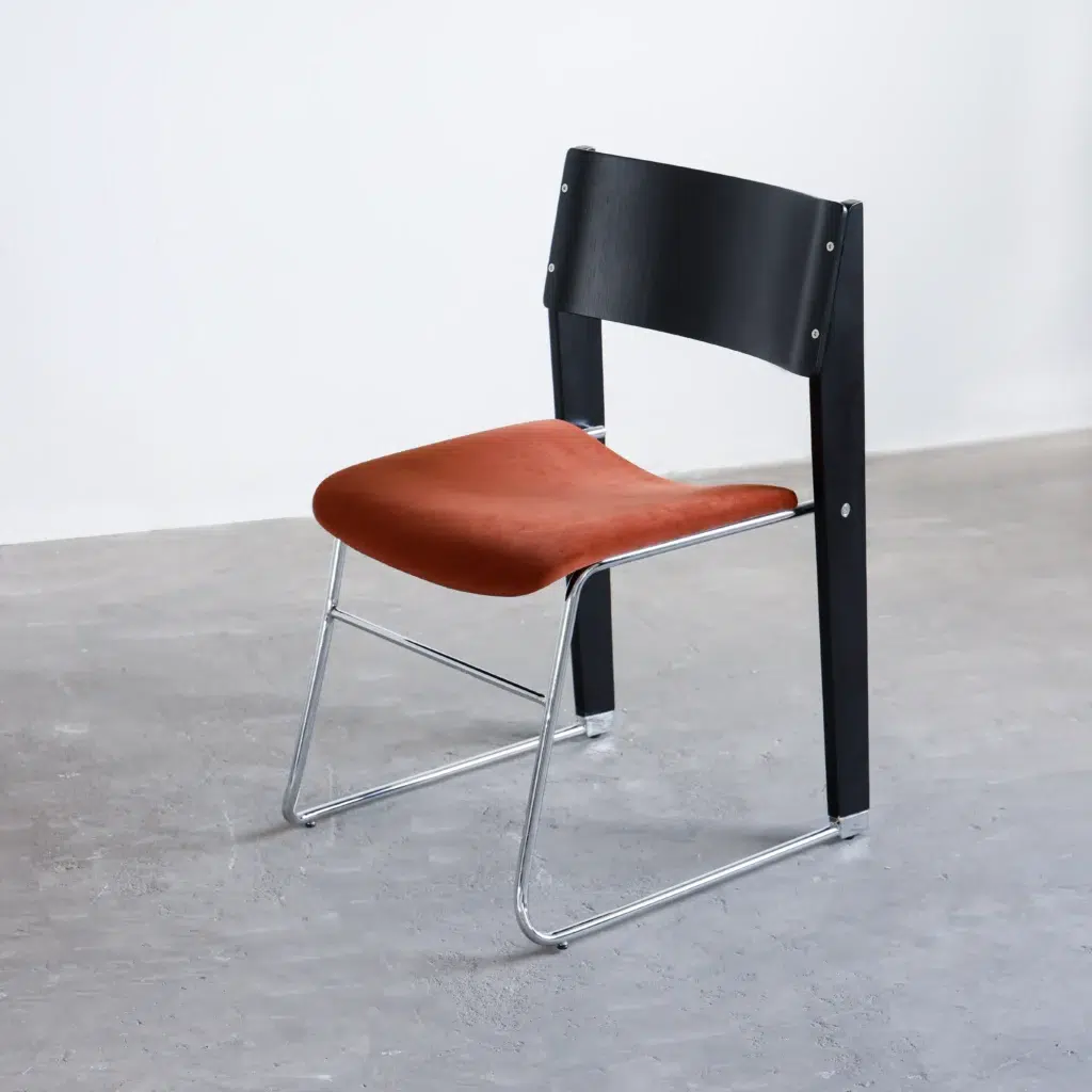A modern chair with a curved black backrest, a rust-coloured cushioned seat, and a chrome metal frame, placed on a plain grey floor against a white wall.