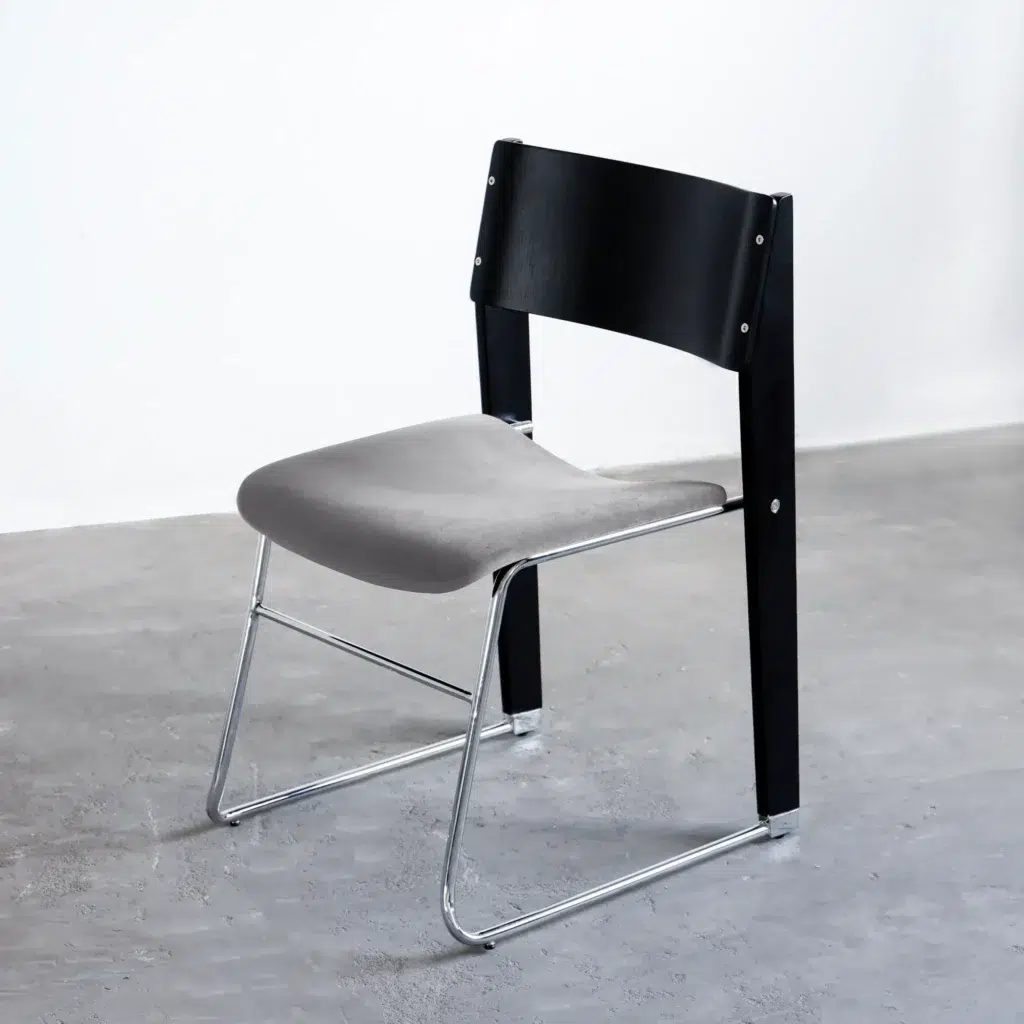 A modern chair with a black wooden backrest, grey cushioned seat, and sleek metal legs, placed on a grey concrete floor against a plain white wall.