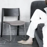 A person dressed in white sits on a grey sofa with their legs extended, one foot on the floor and one foot on the sofa. A modern black and grey chair and a pair of beige slippers are next to them on a dark tiled floor.