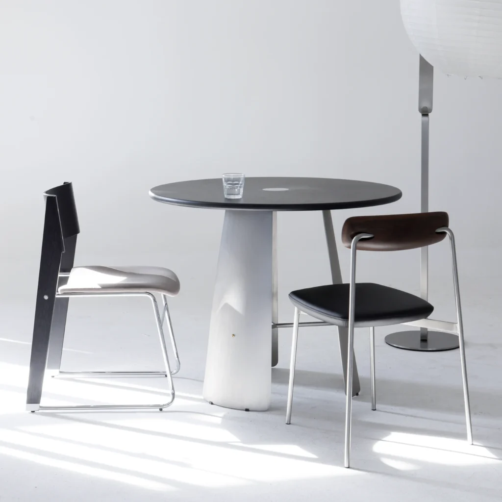 A minimalist set-up with a round black table, two modern chairs, and a clear glass of water. The background is white, and a tall floor lamp stands to the right.