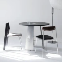 A minimalist set-up with a round black table, two modern chairs, and a clear glass of water. The background is white, and a tall floor lamp stands to the right.