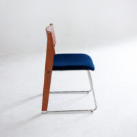 A modern chair with a wooden backrest, blue cushioned seat, and sleek metal legs, photographed from the side against a plain white background.