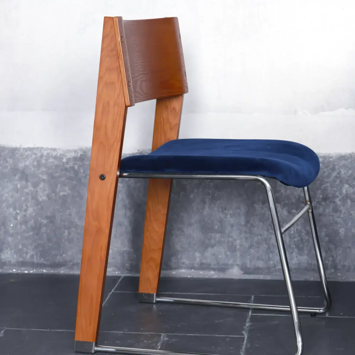Walk Slowly Chair_Brown_Side View A modern chair with a wooden backrest and legs, a blue upholstered seat, and a sleek metal frame, placed on a tiled floor against a grey wall.