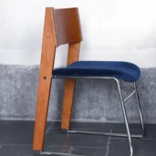 A modern chair with a wooden backrest and legs, a blue upholstered seat, and a sleek metal frame, placed on a tiled floor against a grey wall.