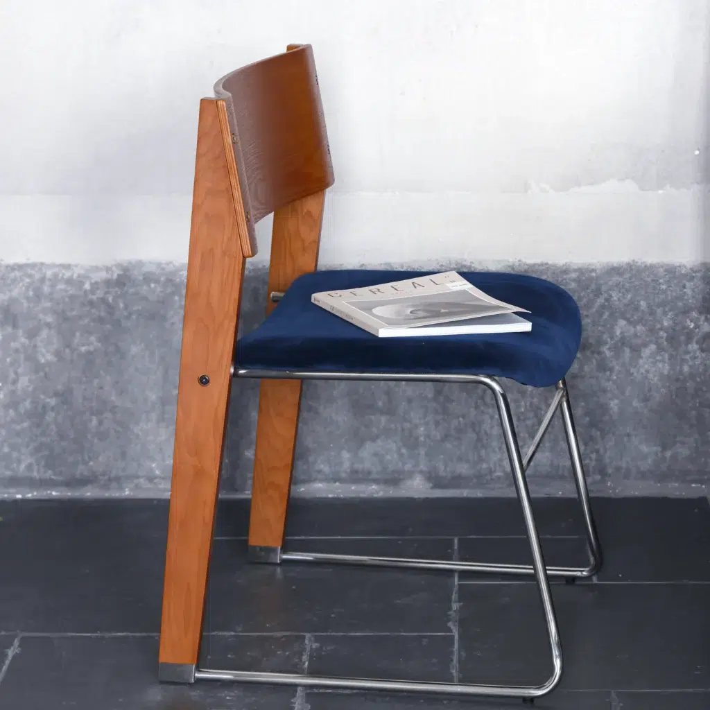 A modern chair with wooden backrest, blue cushioned seat, and metal legs sits on a dark tiled floor. A closed magazine rests on the seat. The background features a textured grey and white wall.