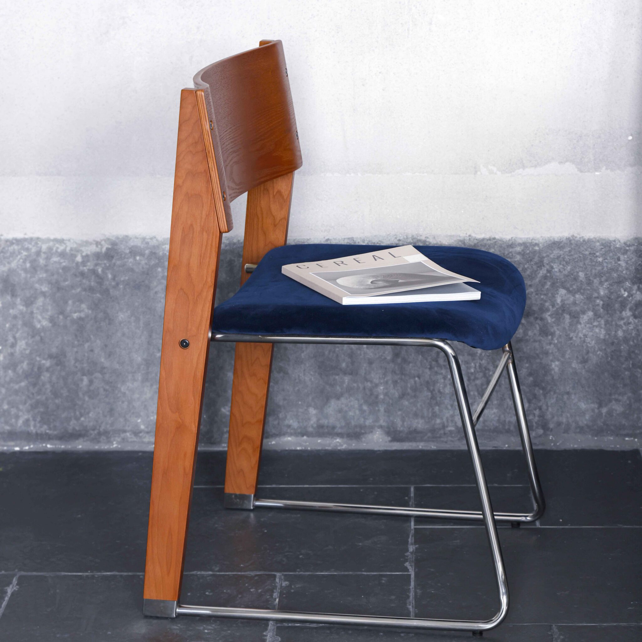 A modern chair with a wooden backrest, blue cushioned seat, and metal legs is placed against a grey wall. A closed magazine or book rests on the seat.