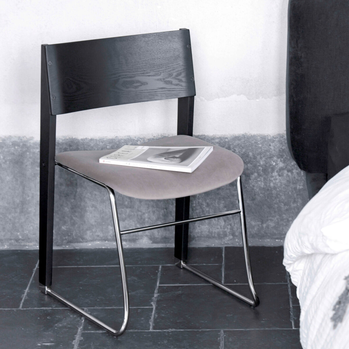 Walk Slowly Chair_Gray_C8 A modern chair with a black wooden backrest, grey cushioned seat, and metal legs, placed on a dark tiled floor next to a bed. A closed book and a magazine lie on the chair’s seat.