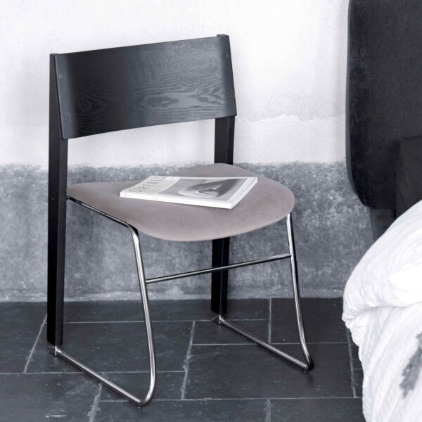 A modern chair with a black wooden backrest, grey cushioned seat, and metal legs, placed on a dark tiled floor next to a bed. A closed book and a magazine lie on the chair’s seat.