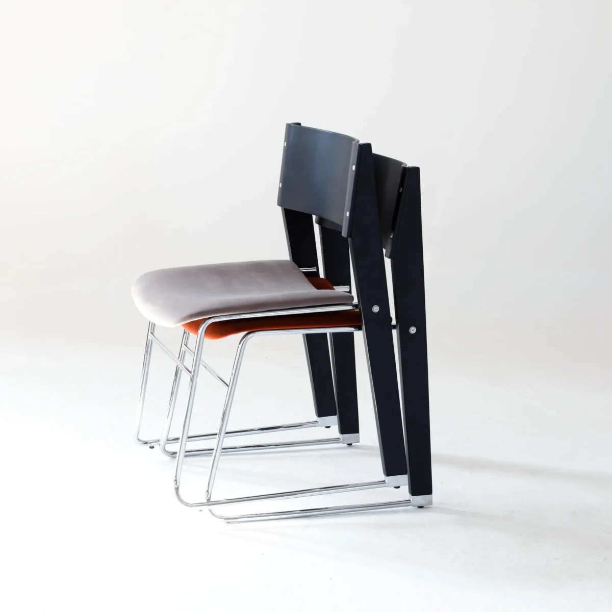 Walk Slowly Chair_Side View_Stack Two modern chairs with metal legs are stacked together. The front chair has a light grey cushioned seat, and the back chair has a dark backrest. Both are placed against a plain white background.