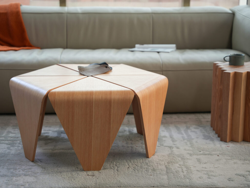 A modern living room features a light wooden geometric coffee table on a grey rug, a beige sofa with an orange throw and books, and a matching side table with a mug on top.