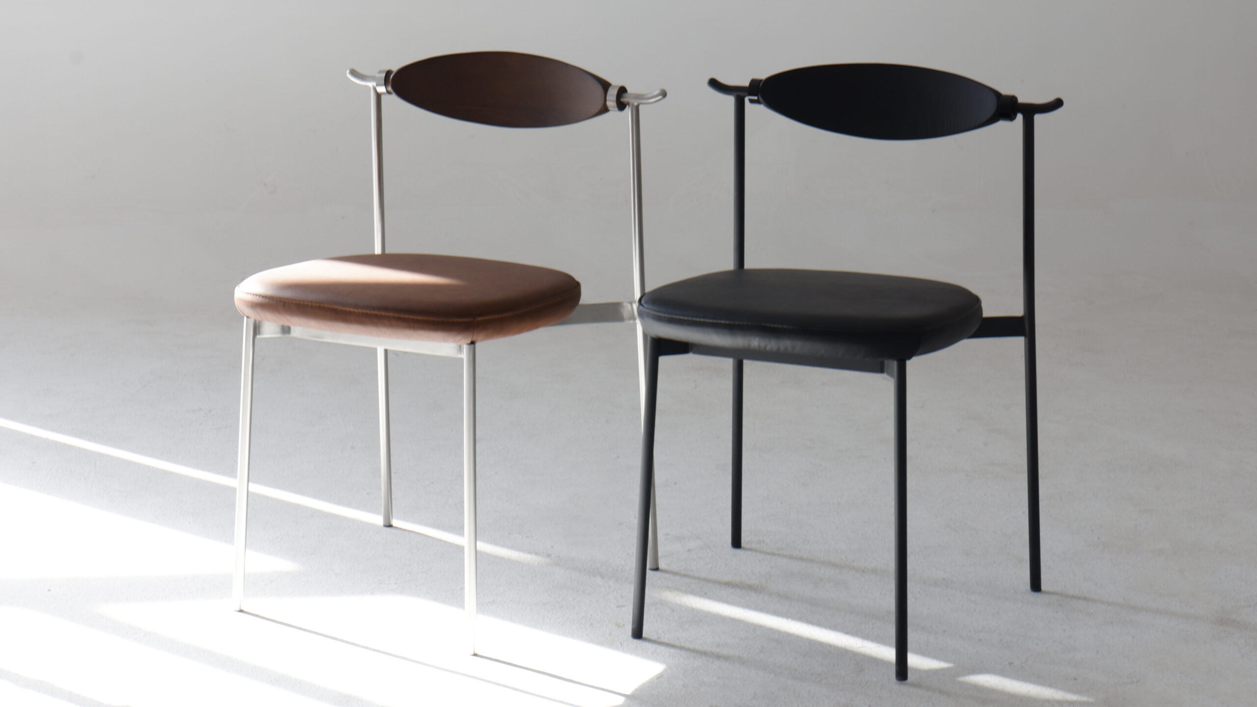 Two modern chairs with curved backrests and padded seats are placed side by side on a light floor. One chair is brown with silver legs; the other is black with matching black legs. Sunlight streams across the scene.