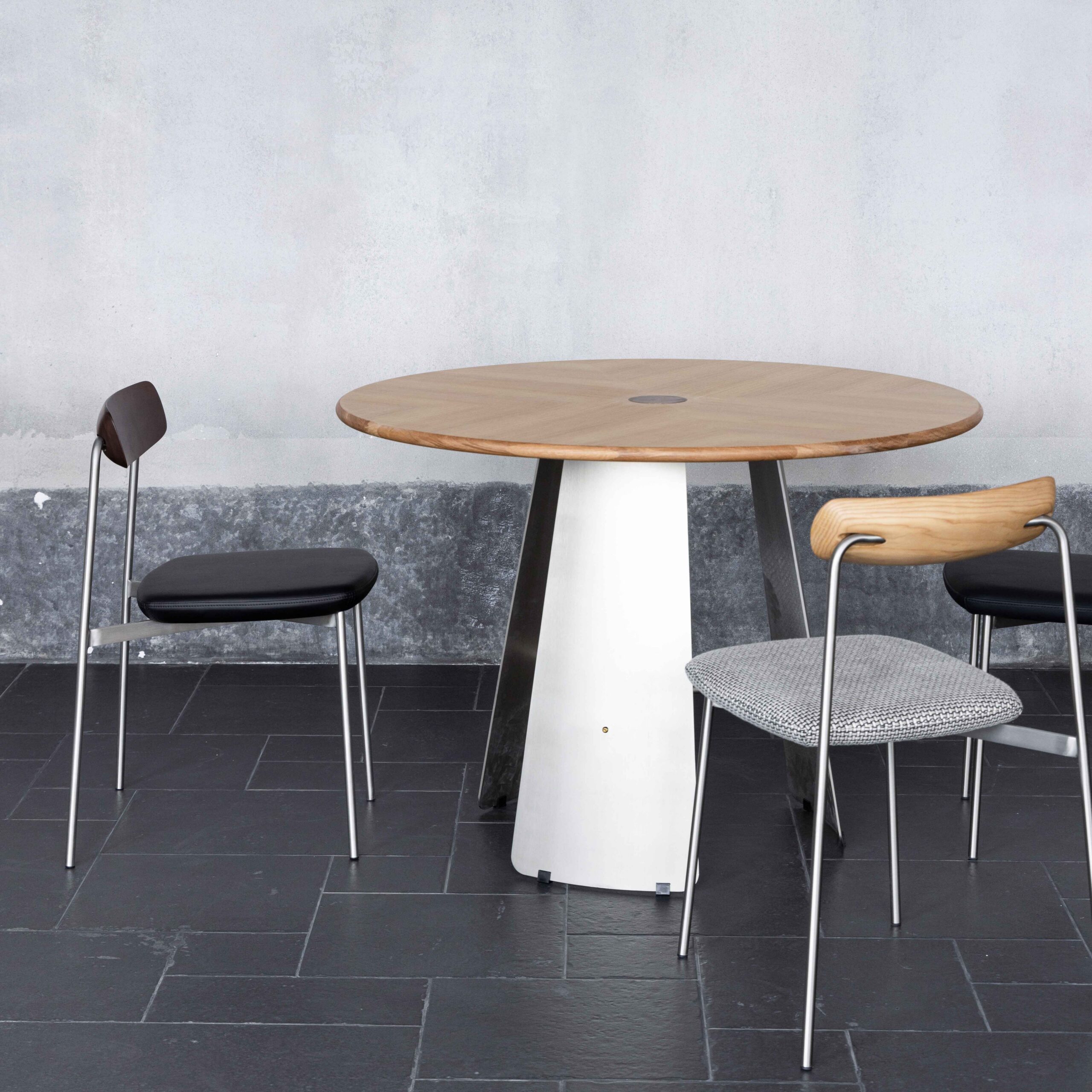 A round wooden table with a white and metal base stands on a black tiled floor, accompanied by two minimalist chairs with metal legs and different seat and backrest designs, against a light grey wall.