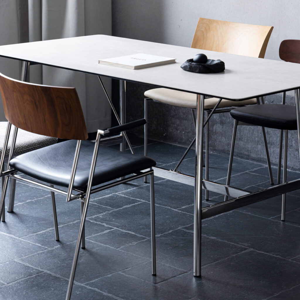 A modern dining area with a rectangular table featuring a sleek metal frame, surrounded by three contemporary chairs in wood, black, and beige. A closed book and computer mouse rest on the tabletop.