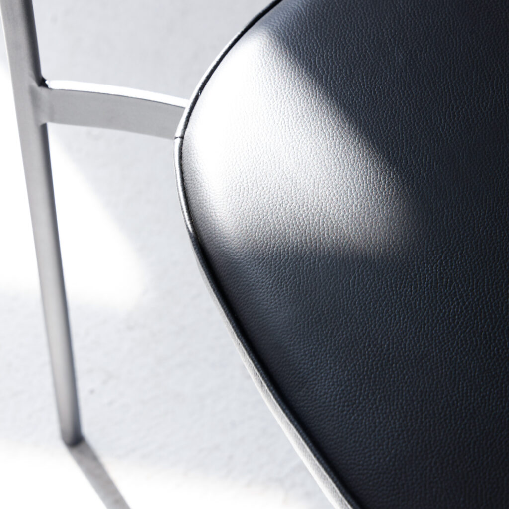 Close-up of a modern chair with a curved, black leather seat and a metal frame, with sunlight casting soft shadows on the seat and white floor.