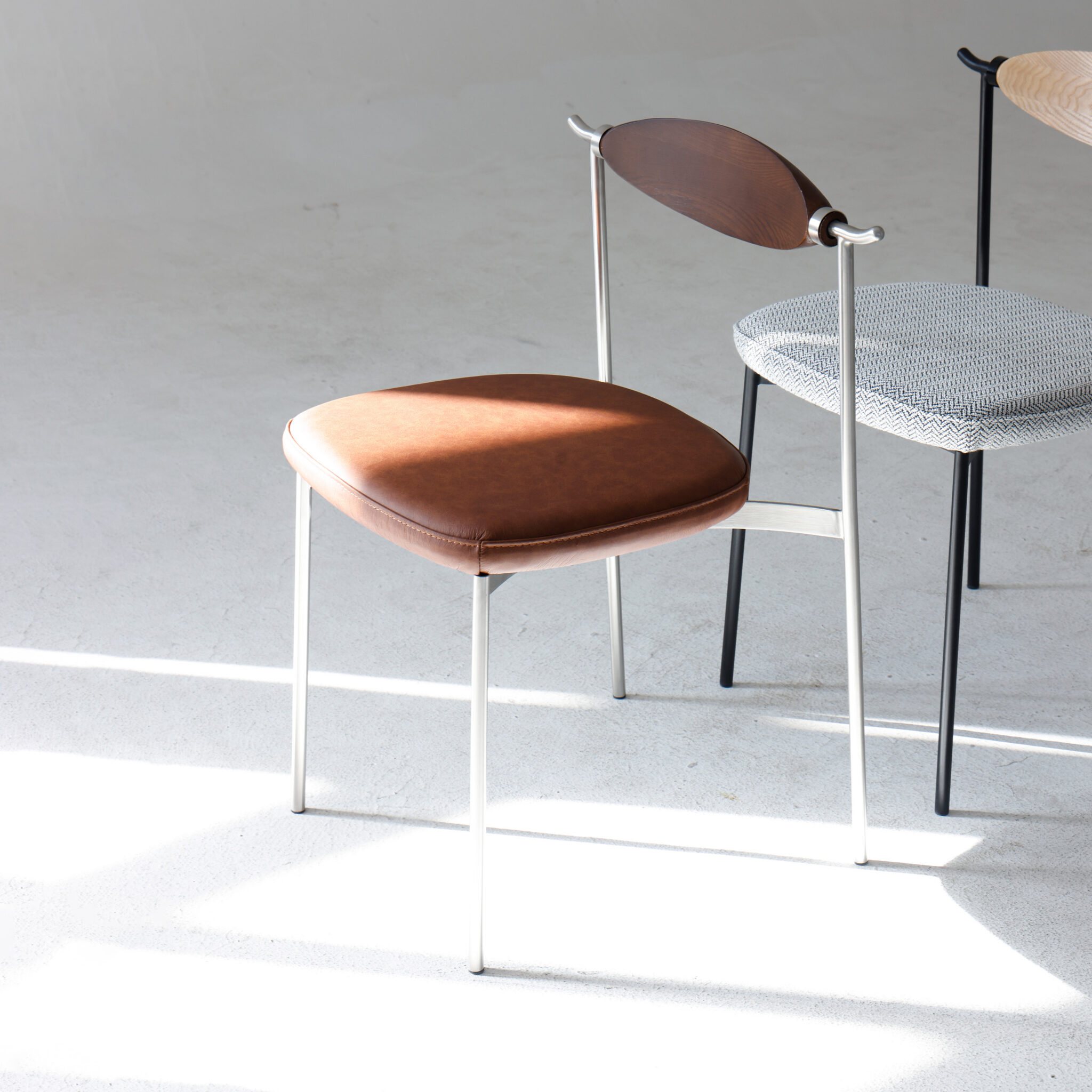 A modern chair with a brown leather seat, metal legs, and a curved wooden backrest sits on a light floor. Behind it is another chair with a light grey upholstered seat and black metal legs, partially visible. Sunlight streams across the floor.