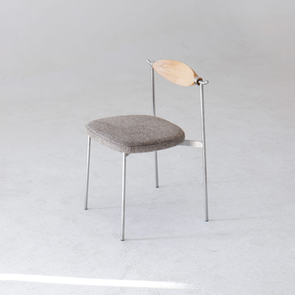 A minimalist chair with thin metal legs, a grey upholstered seat, and a light wooden oval backrest, set against a plain, light background.