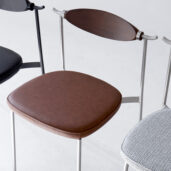 Three modern chairs with metal frames are shown. One has a brown leather seat and wooden backrest, another has a black seat and back, and the third has a light grey patterned seat. All are placed on a light-coloured floor.