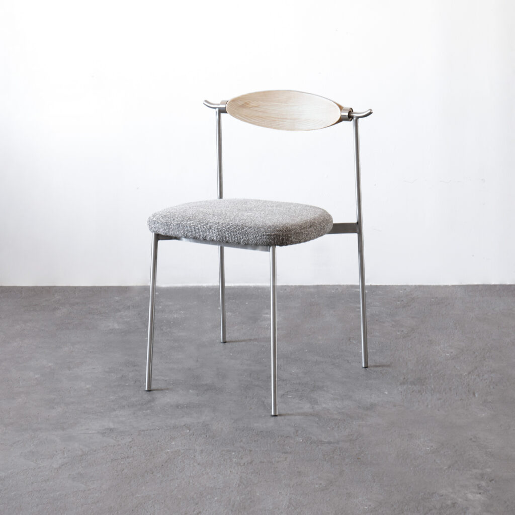 A minimalist chair with thin metal legs, a round grey cushioned seat, and a light wooden curved backrest, set against a plain white wall and grey concrete floor.
