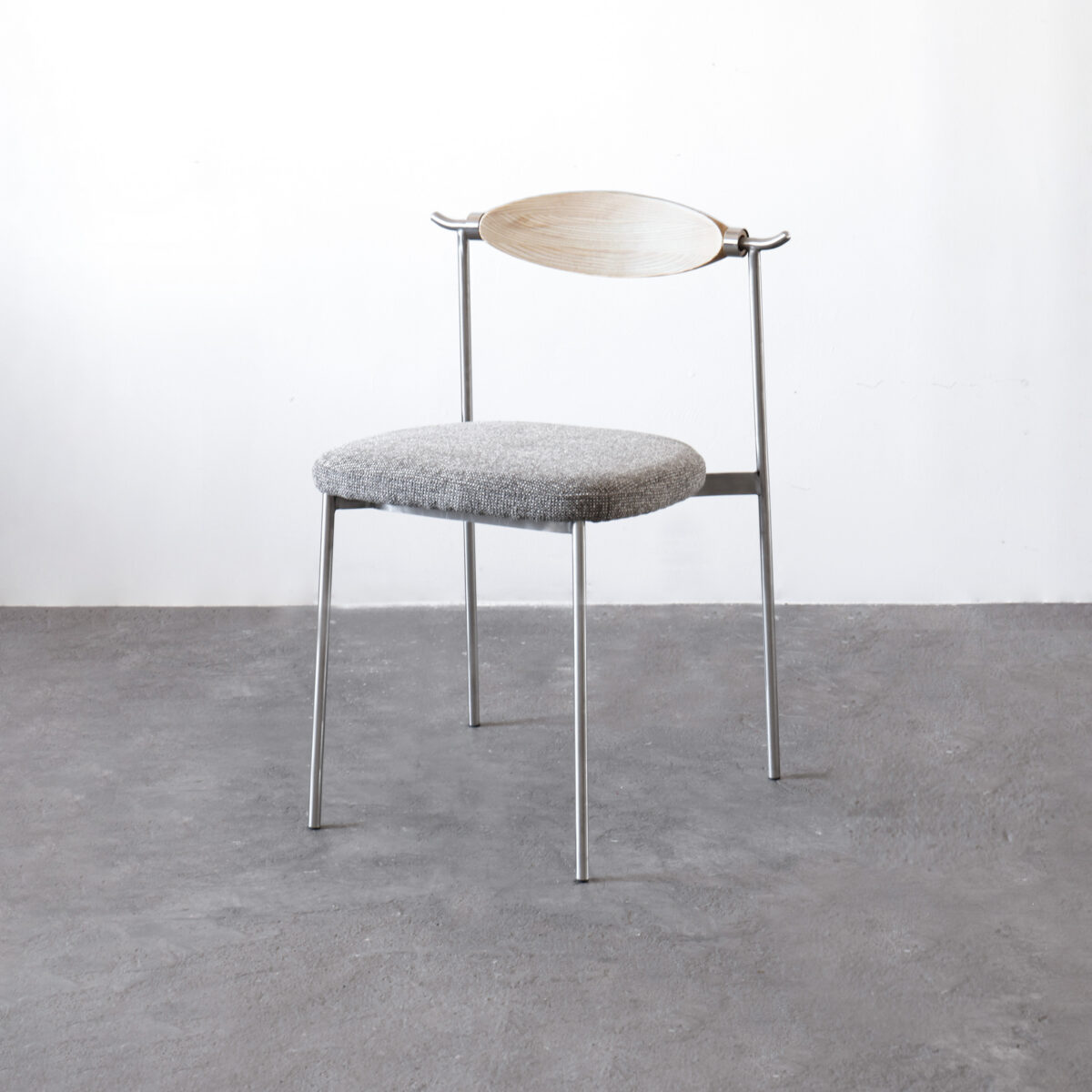 05_ASSC-CR-ASNA-01 A minimalist chair with thin metal legs, a round grey cushioned seat, and a light wooden curved backrest, set against a plain white wall and grey concrete floor.