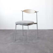 A minimalist chair with thin metal legs, a round grey cushioned seat, and a light wooden curved backrest, set against a plain white wall and grey concrete floor.