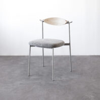 A minimalist chair with thin metal legs, a round grey cushioned seat, and a light wooden curved backrest, set against a plain white wall and grey concrete floor.