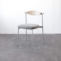 A minimalist chair with thin metal legs, a round grey cushioned seat, and a light wooden curved backrest, set against a plain white wall and grey concrete floor.