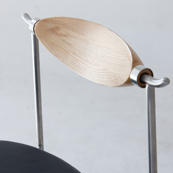A modern chair with a curved, light wood backrest attached to a metal frame, and a dark upholstered seat partially visible at the bottom left. The design is minimalist and contemporary.